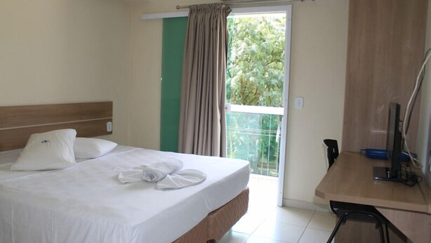 A hotel room with a bed, a desk, and a view of greenery outside. - Hotel do Farol (Cubatao)