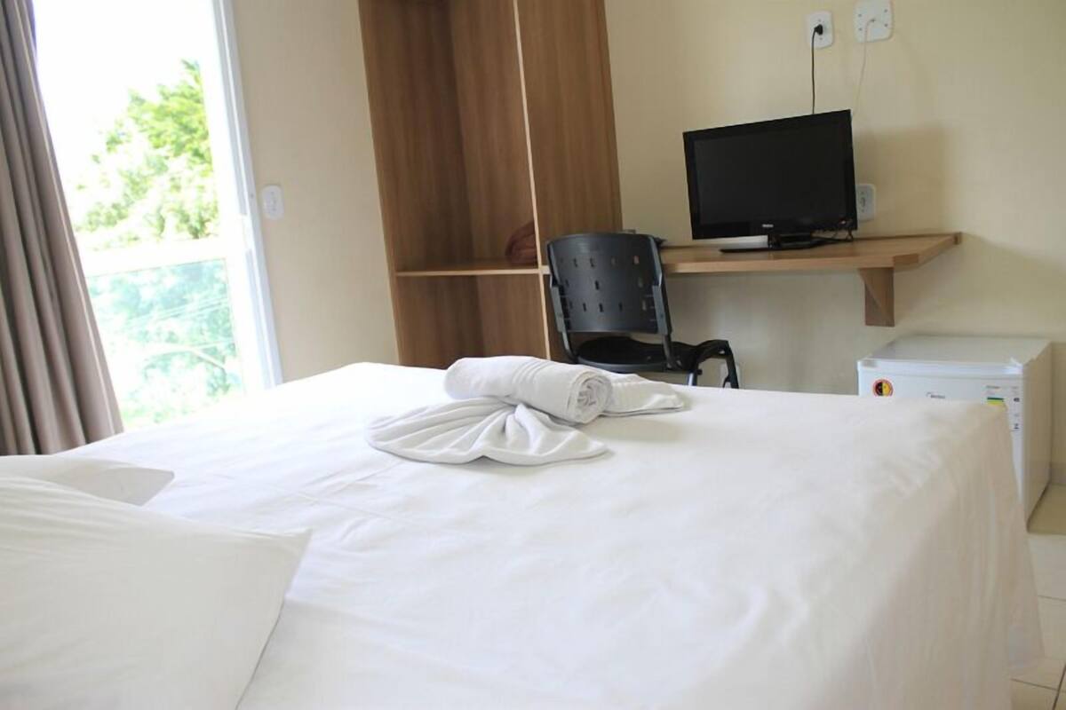 A hotel room with a bed, a desk with a computer, a chair, and a small white appliance.