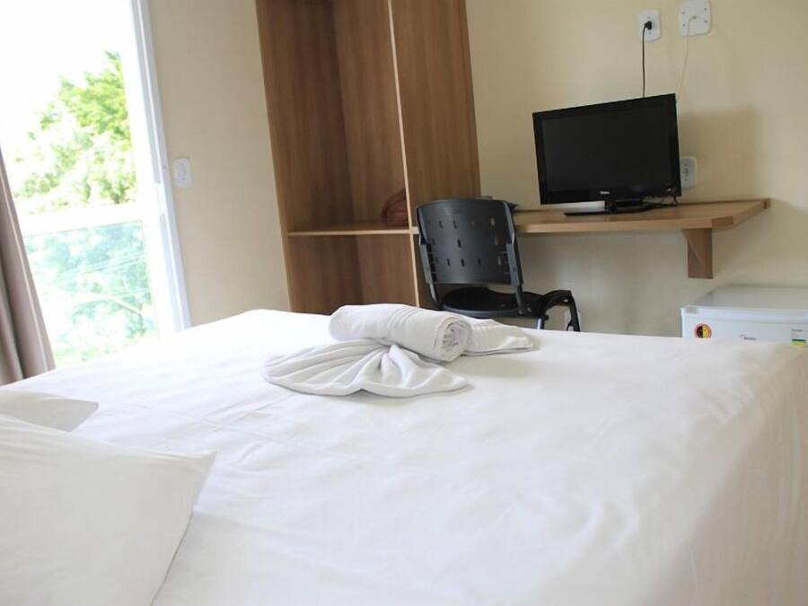 Una habitación de hotel con una cama, un escritorio con una computadora, una silla y un electrodoméstico blanco pequeño.