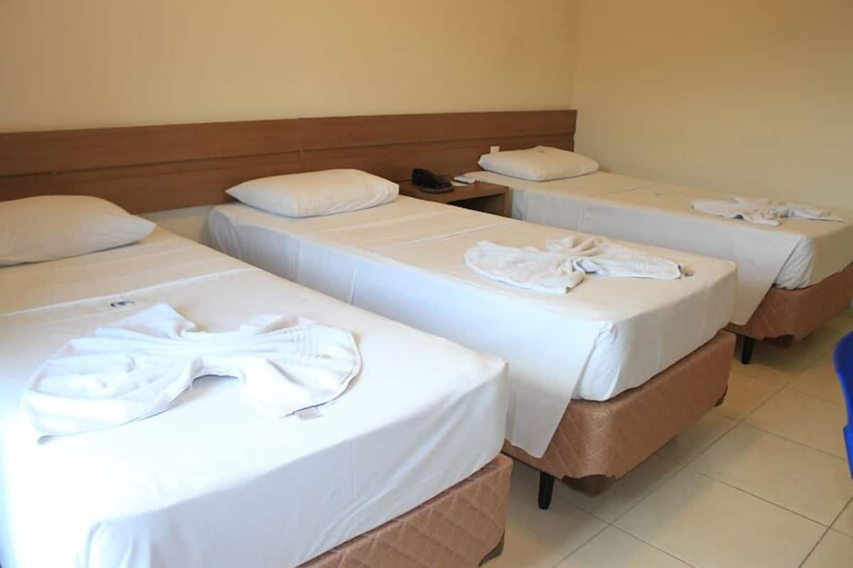 A hotel room with three single beds, each with a white bedspread and pillow. There is a wooden headboard, a nightstand with a phone, and a blue chair.
