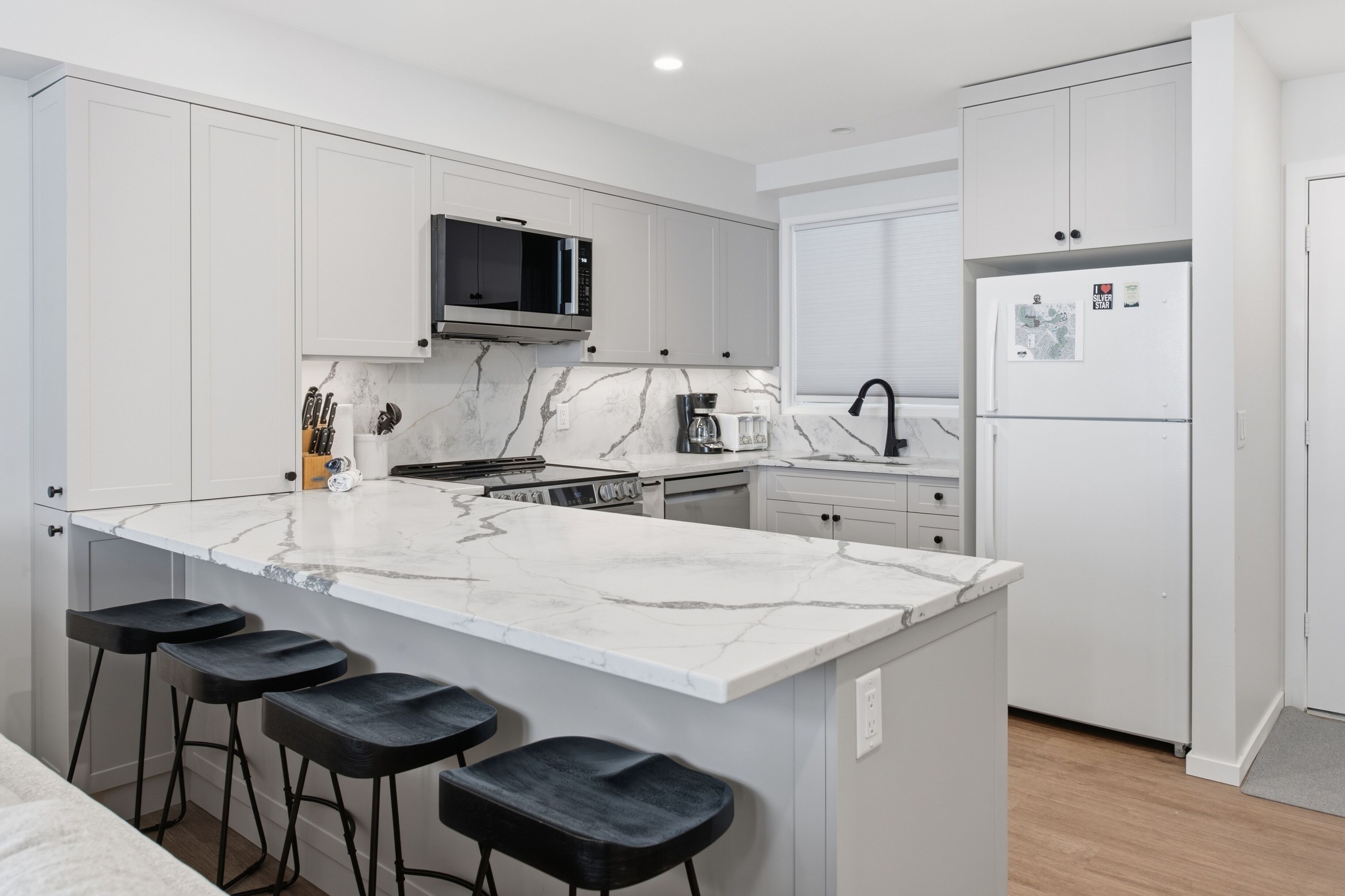 A modern kitchen with white cabinets, a marble countertop, a built-in microwave, a refrigerator, and bar stools.