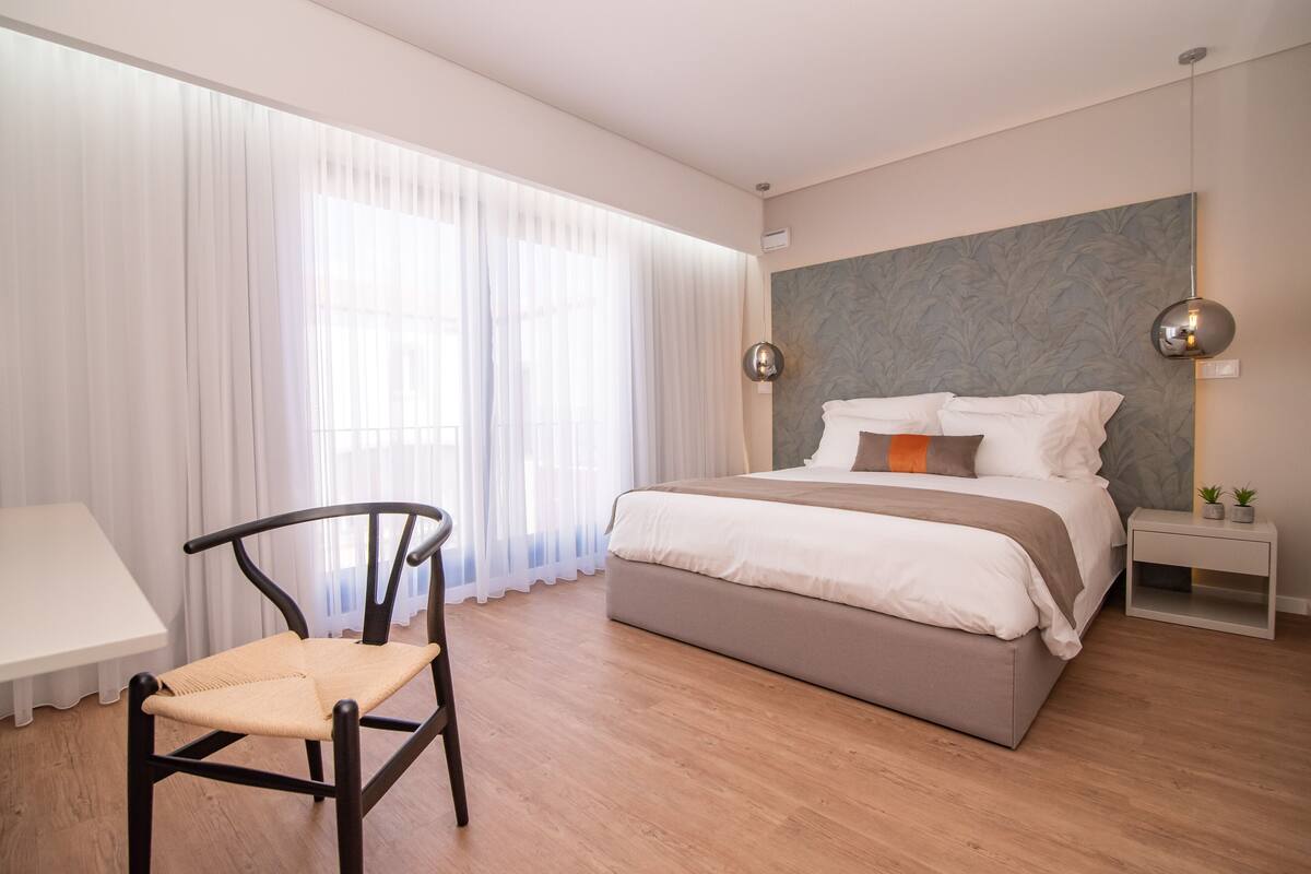 Un dormitorio moderno con una cama, una silla, una mesita de noche y una ventana con cortinas translúcidas.