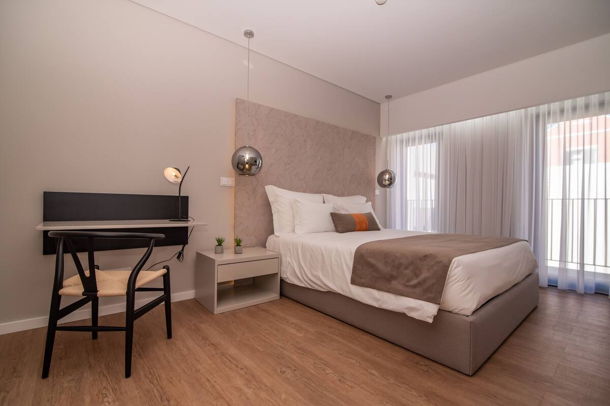 Un dormitorio moderno con cama, mesita de noche, silla, escritorio y ventanas amplias.