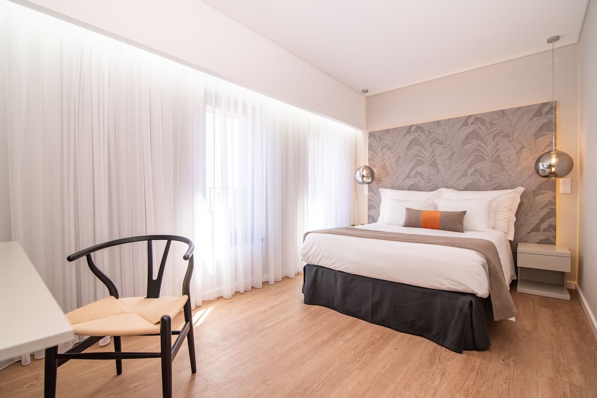 Un dormitorio moderno con cama, una silla, una mesita de noche y un amplio ventanal con cortinas translúcidas.
