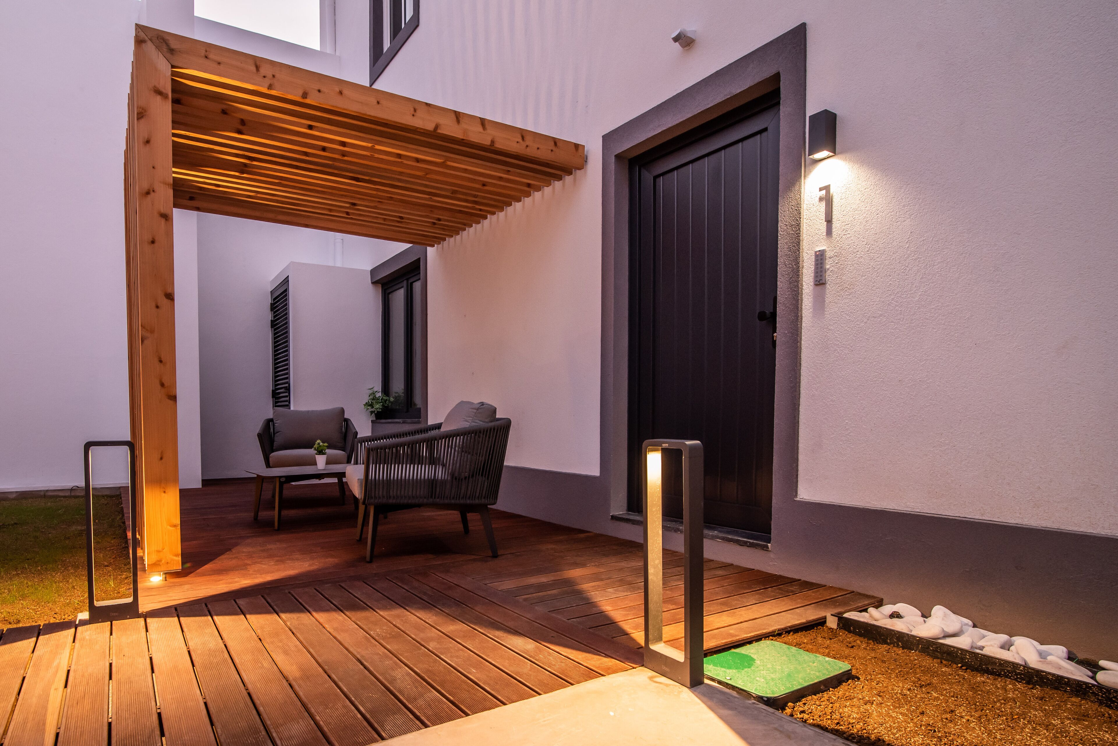 A modern outdoor patio with wooden flooring, a pergola, a seating area with a chair and table, and a door with a number plaque.