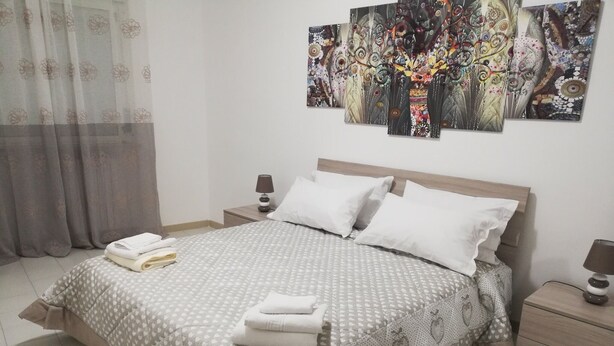 A neatly made bed with a patterned bedspread, two bedside lamps, and a wall with abstract art. - Madrigio ... at home (Casamassima)