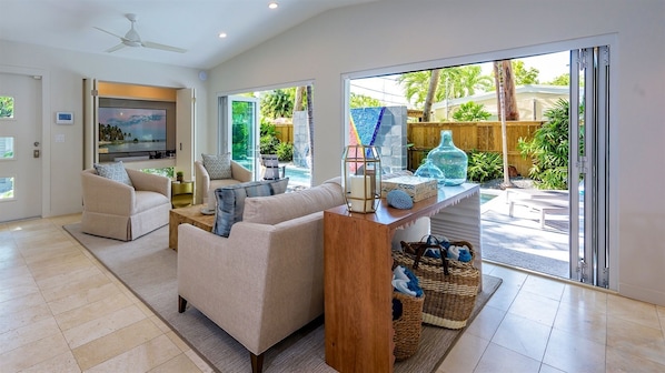 A spacious living room with a sofa, a coffee table, and a TV. There is a sliding glass door leading to a patio with a pool and greenery.