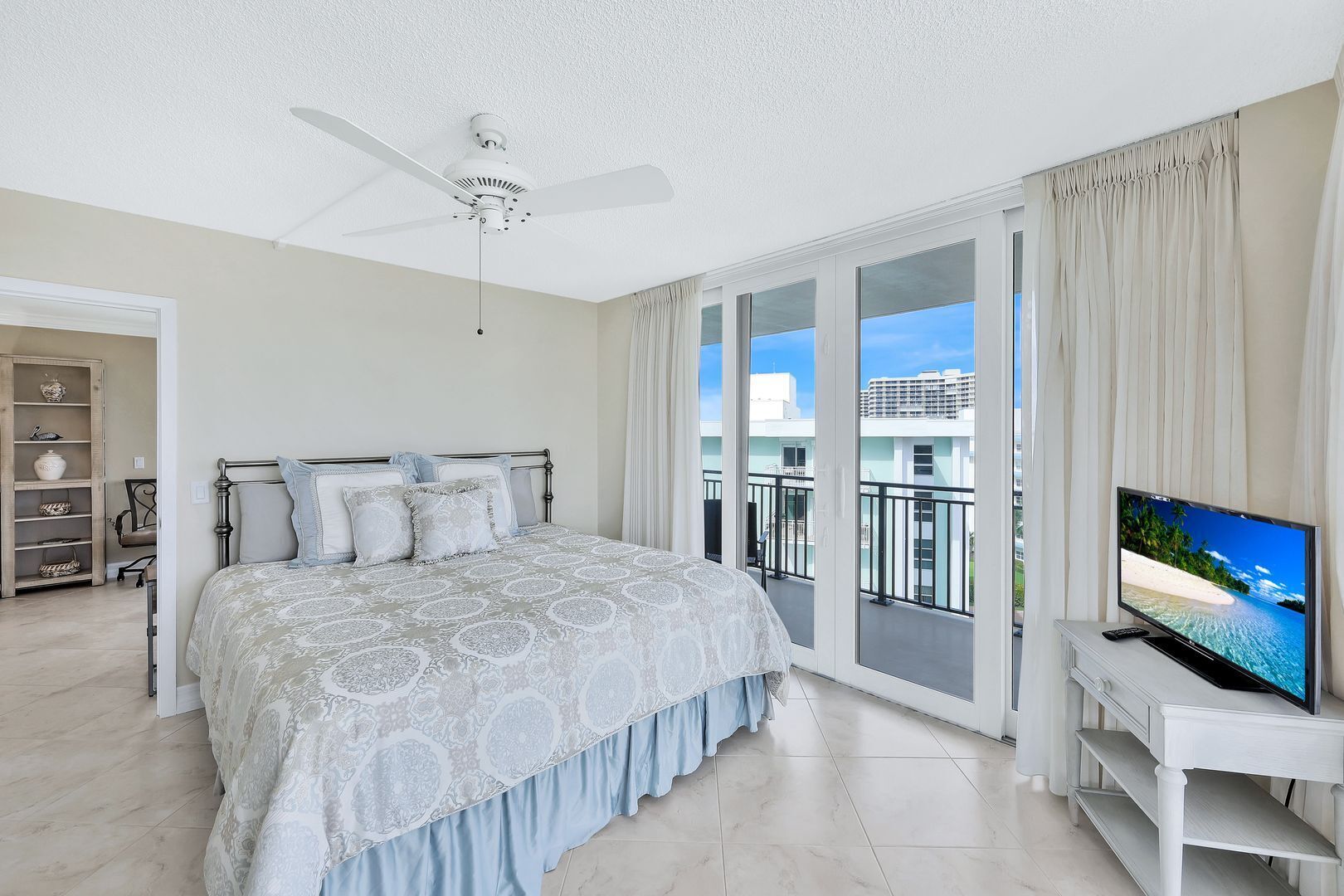 A bedroom with a bed, a ceiling fan, a TV, and a balcony with a view.