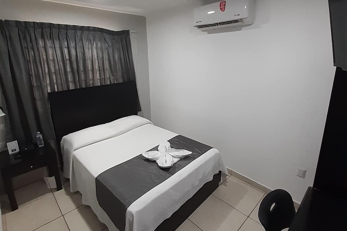 A hotel room with a bed, a desk, a chair, an air conditioning unit, and a television.