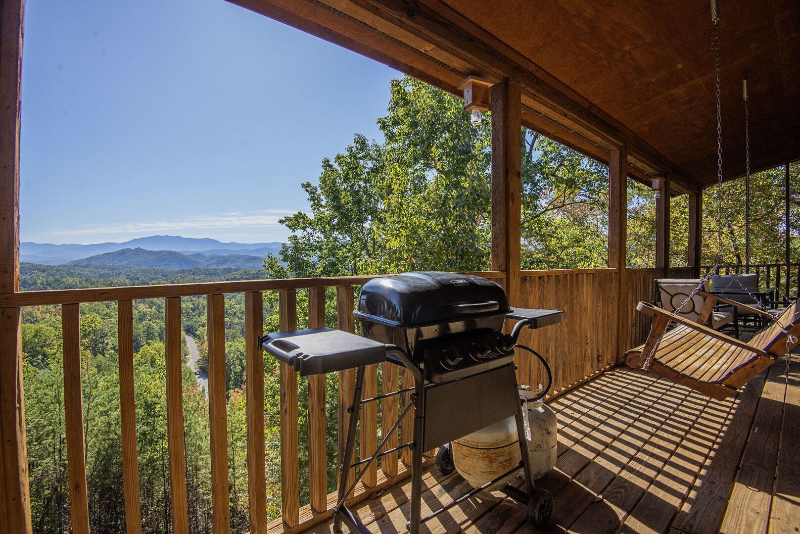 A covered deck with a grill and seating area, overlooking a forested landscape with mountains in the distance.