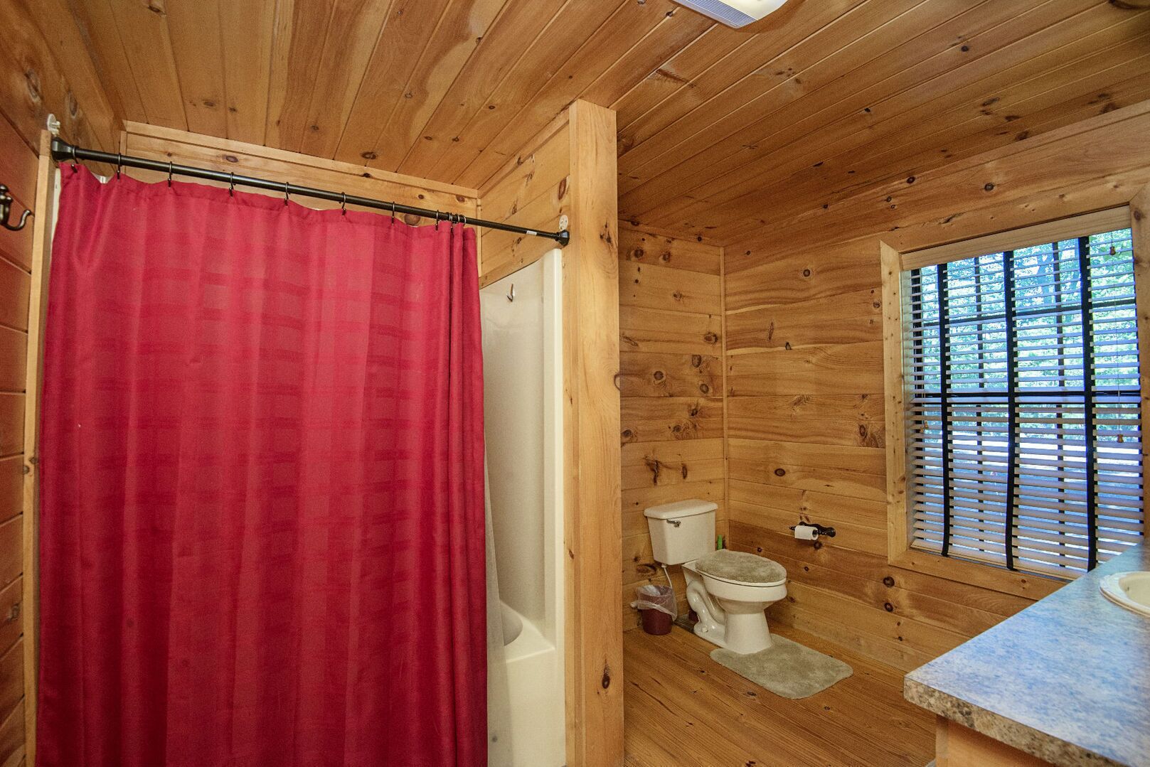 A wooden cabin bathroom with a red shower curtain, a toilet, and a window with blinds.