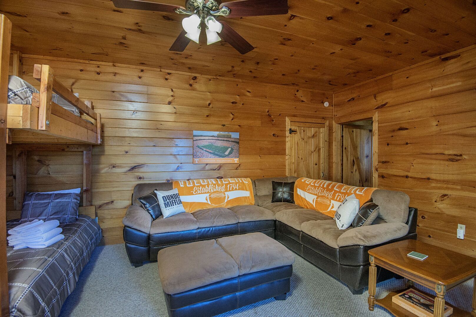 A wooden cabin interior with a sofa, ottoman, and a bed with folded towels.