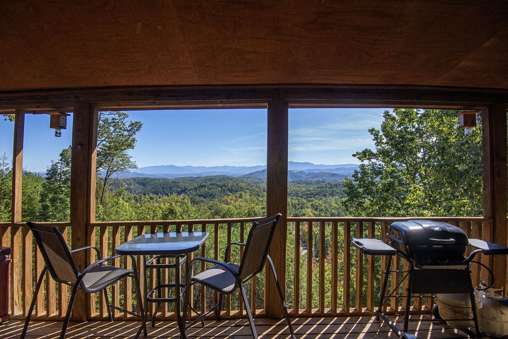 A wooden deck with a view of a forest and mountains. There are three tables and two chairs, and a grill is visible on the right.