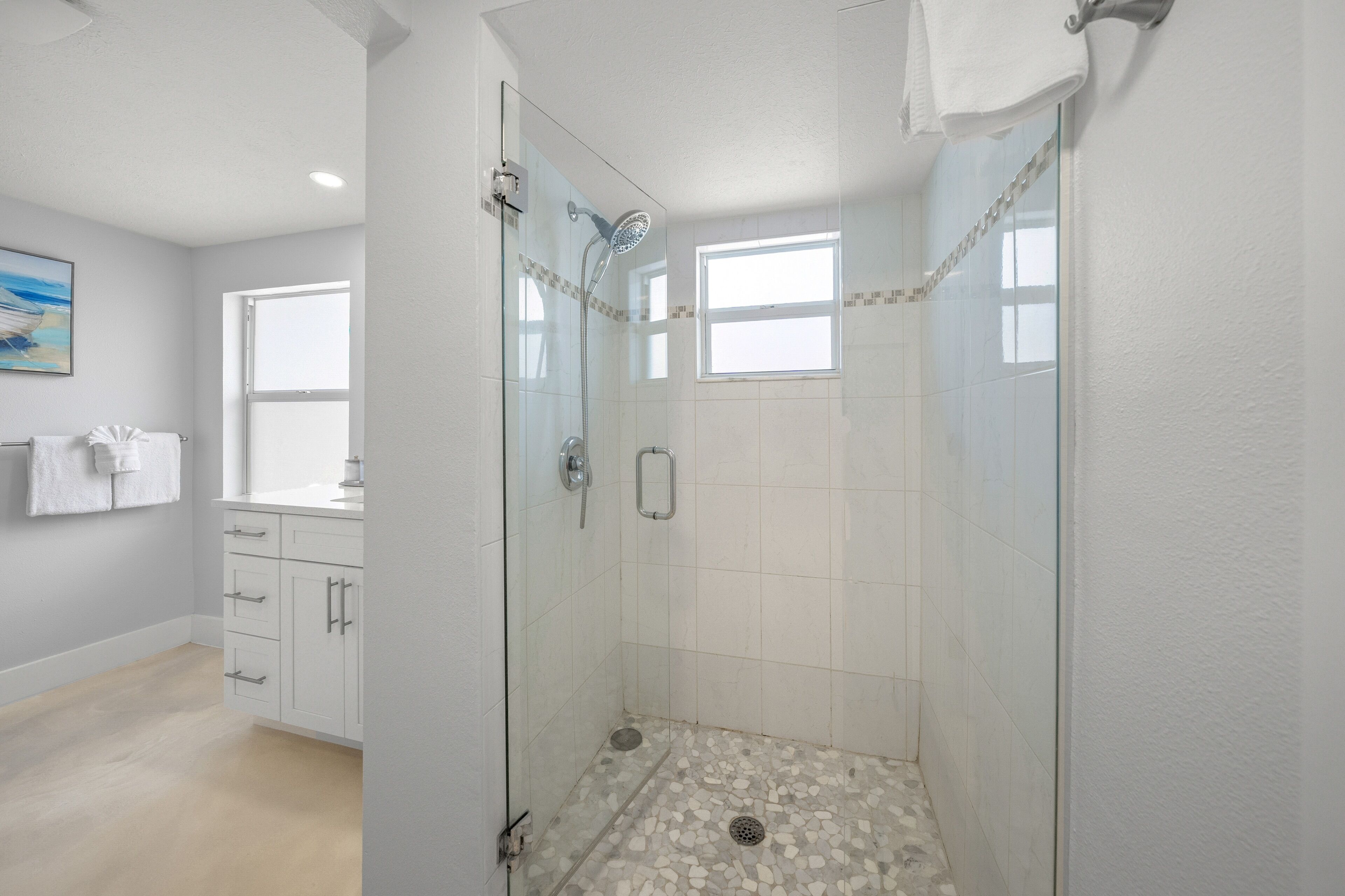 A modern bathroom with a glass-enclosed shower, white tiles, and a window providing natural light.