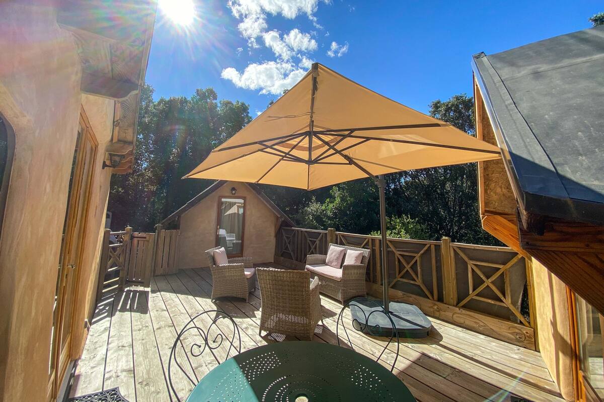Eine Holzterrasse mit Gartenmöbeln, einem großen Sonnenschirm und einem kleinen Gebäude mit einer Tür.