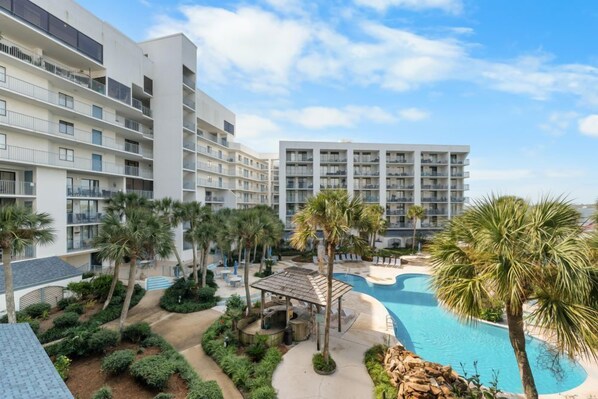 A hotel with multiple floors, outdoor swimming pool, palm trees, and a covered seating area.