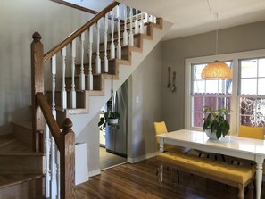 A staircase with wooden steps and white balusters, a dining area with a white table and bench, and a window with a hanging light fixture.