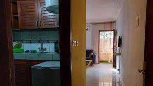 Una cocina con armarios y un refrigerador, un área de estar con sofá y televisor, y una puerta que da a otra habitación.