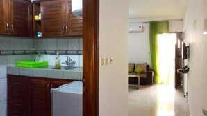 Una cocina con gabinetes de madera y un refrigerador, una sala de estar con un sofá y una mesa de centro, y un pasillo con una puerta que da a otra habitación.