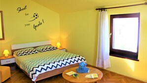 Una cama bien hecha con una colcha estampada, una mesita de noche con una lámpara, una pequeña mesa redonda con un globo terráqueo y libros, y una ventana con cortinas.