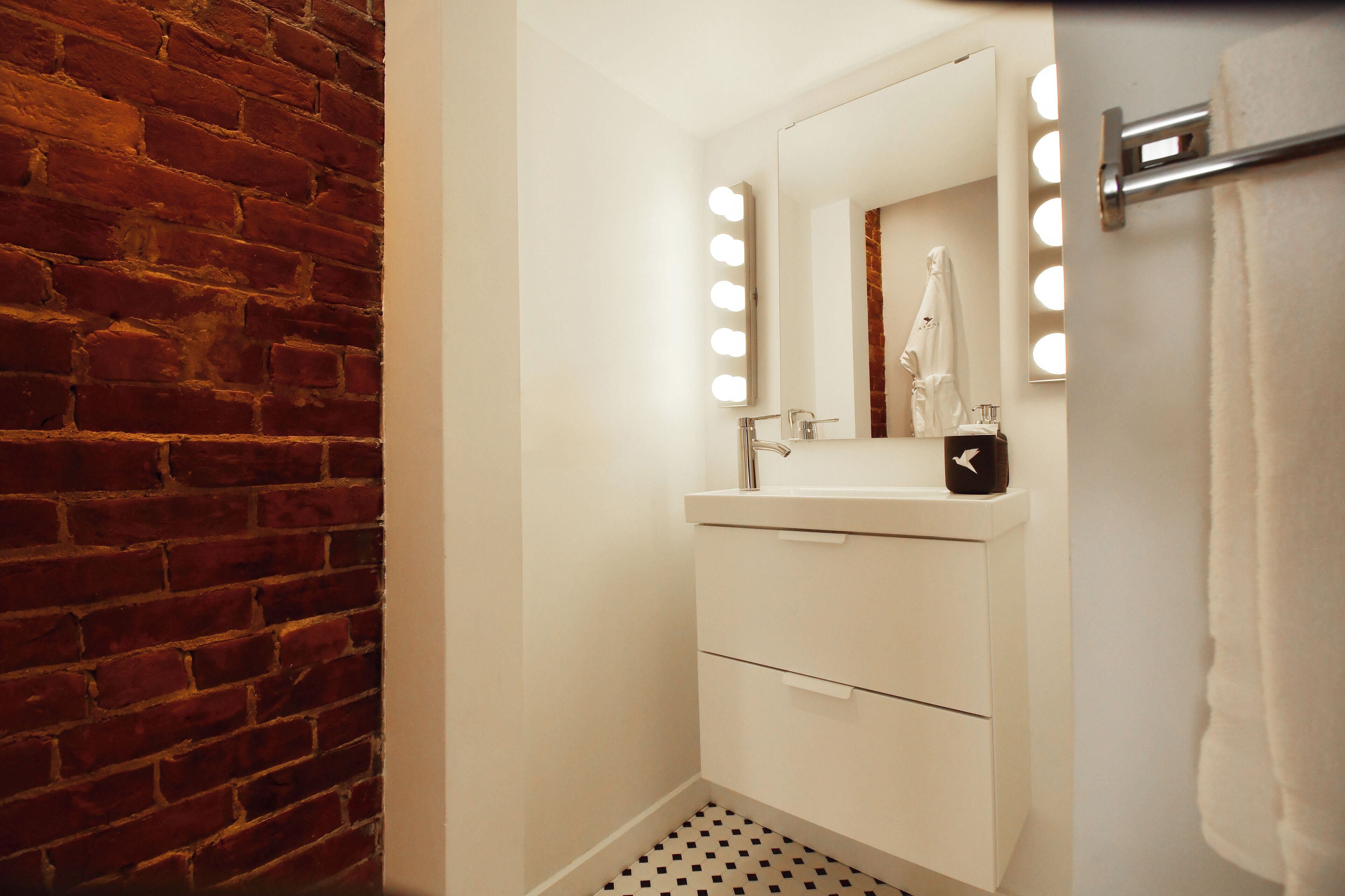 Un baño con pared de ladrillo, un lavamanos blanco con espejo y un perchero para toallas.