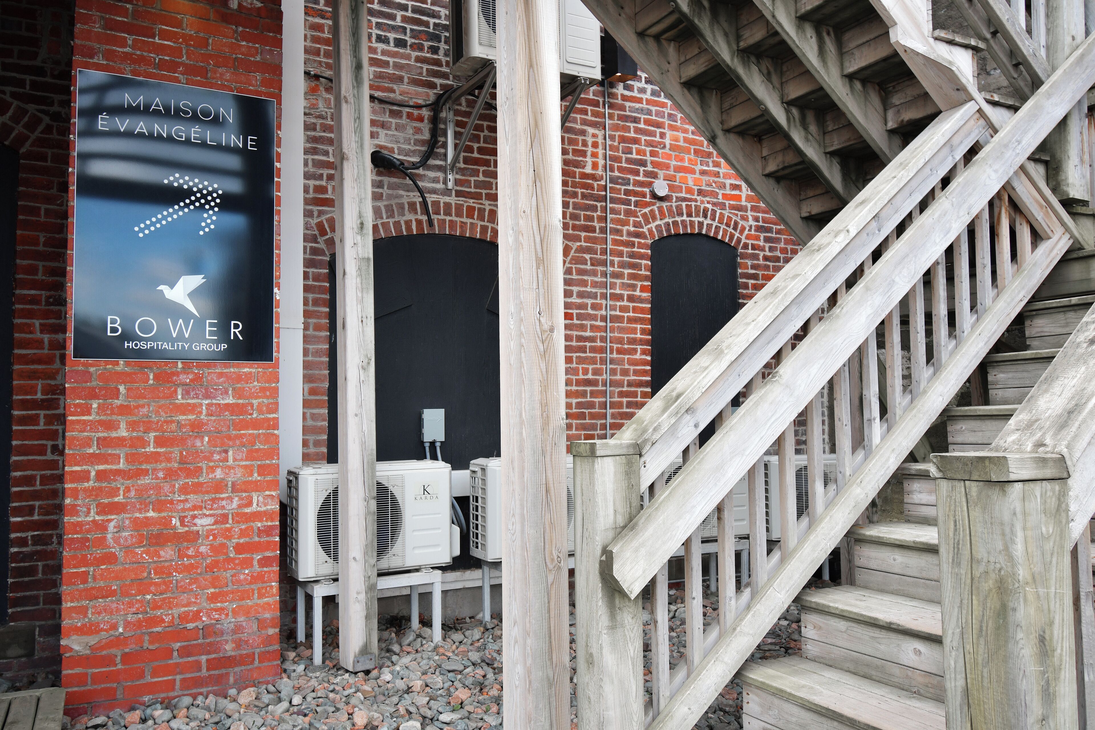A brick building with a sign for Maison Evangeline Bower Hospitality Group, outdoor air conditioning units, and a wooden staircase.
