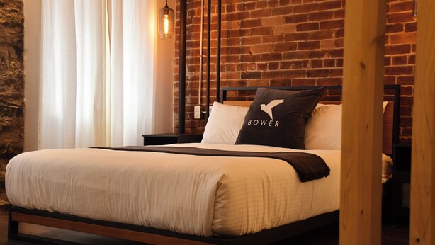 Una cama con estructura de madera, ropa de cama blanca y una almohada con la palabra "BOWER".