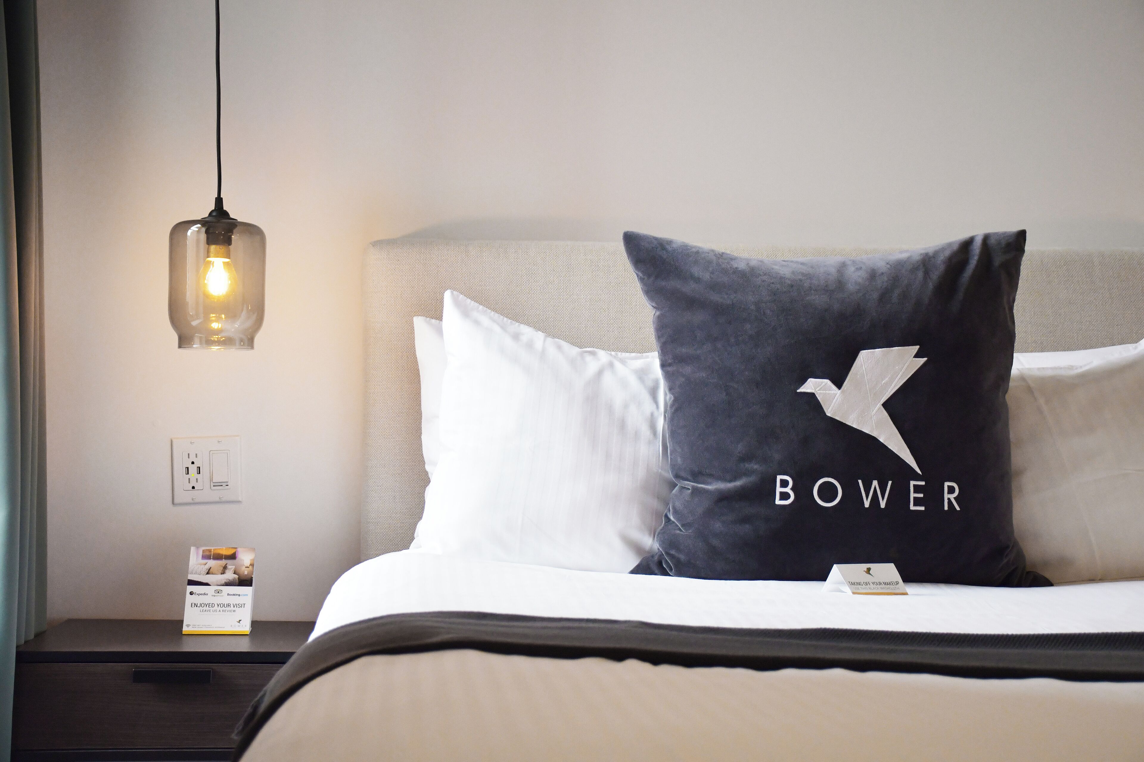 Una cama bien hecha con almohadas blancas y gris oscuro, una lámpara colgante, una mesita de noche con toma de corriente y una pequeña señalización sobre la cama.