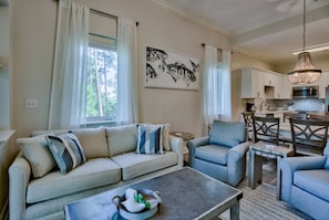 A living room with a sofa, chairs, a coffee table, and a dining area with chairs and a table. There is a kitchen visible in the background.
