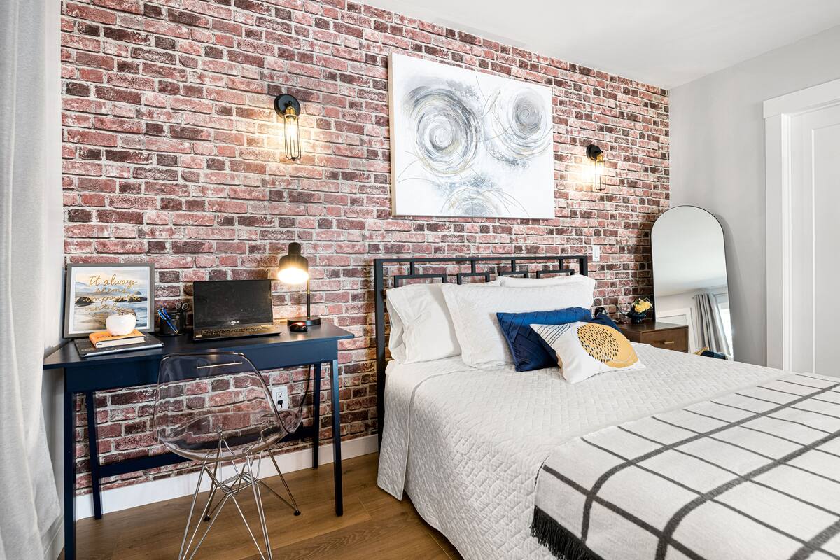 Un dormitorio con pared de ladrillo, cama con ropa de cama blanca, escritorio con una computadora portátil y una silla.