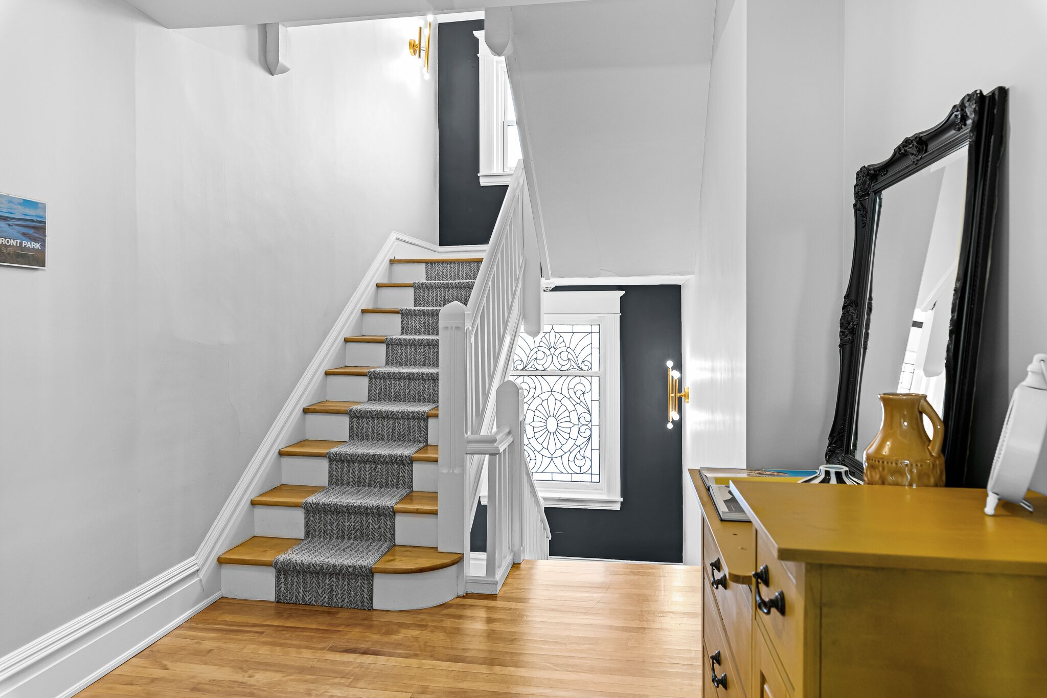 Een trap met houten treden en een patroon vloerkleed, een witte leuning, een donkere deur met decoratief raam, en een dressoir met spiegel en vaas.