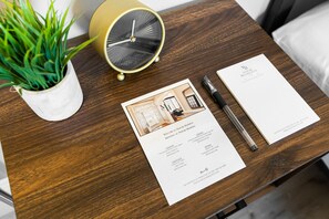 A wooden table with a white potted plant, a vintage-style alarm clock, a business card, and a printed document with a building image and text. - Manoir Highfield (Moncton)