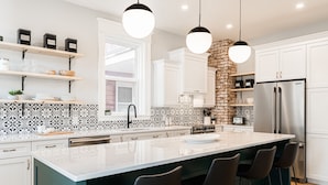 A modern kitchen with white cabinets, a large island with bar stools, a stainless steel refrigerator, and pendant lighting. - Manoir Highfield (Moncton)