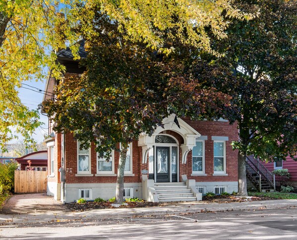 A two-story brick house with a front porch, a small front yard, and a wooden fence. - Manoir Highfield (Moncton)