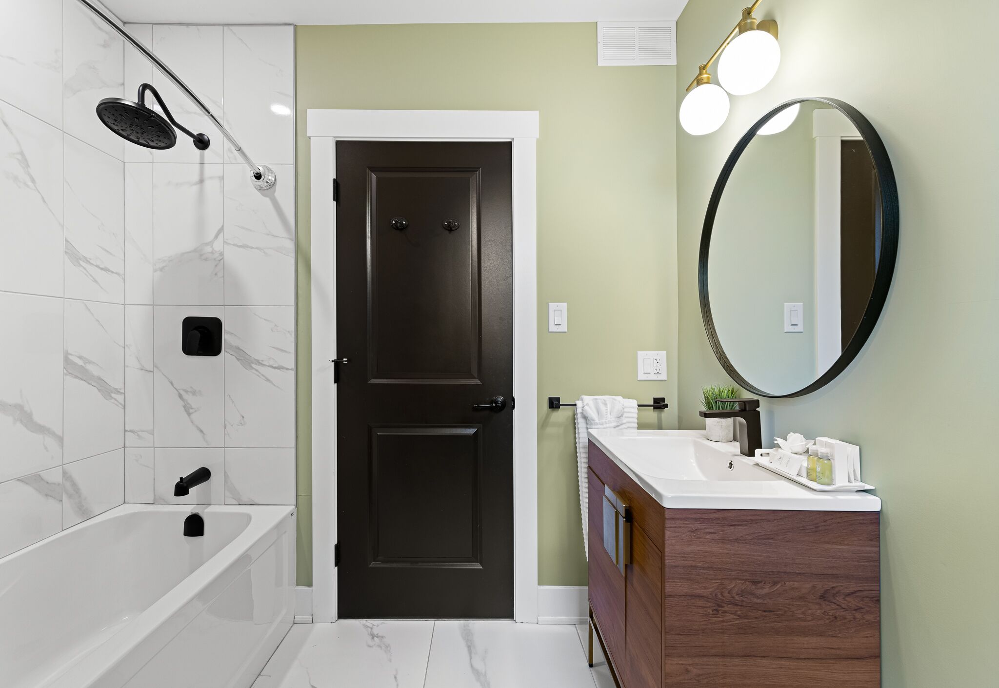 A bathroom with a bathtub, a shower, a dark door, a vanity with a mirror, and a light fixture.
