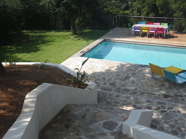 Piscina com deck de pedra, rodeada por gramado e árvores. Há uma mesa com cadeiras coloridas e uma pequena área de jardim com plantas.