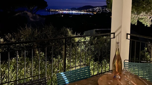 Ein Balkon mit Holztisch, einer Flasche und Gläsern, mit Blick auf eine nächtliche Stadtlandschaft.
