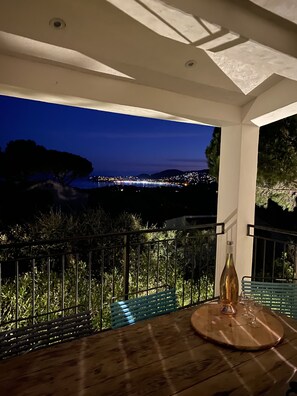 Varanda com mesa de madeira, uma garrafa e copos, com vista para a cidade à noite.