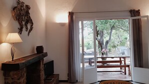 Et hyggeligt værelse med en stenvægspejs, en lampe og udsigt til en udendørs terrasse med et bord og stole.