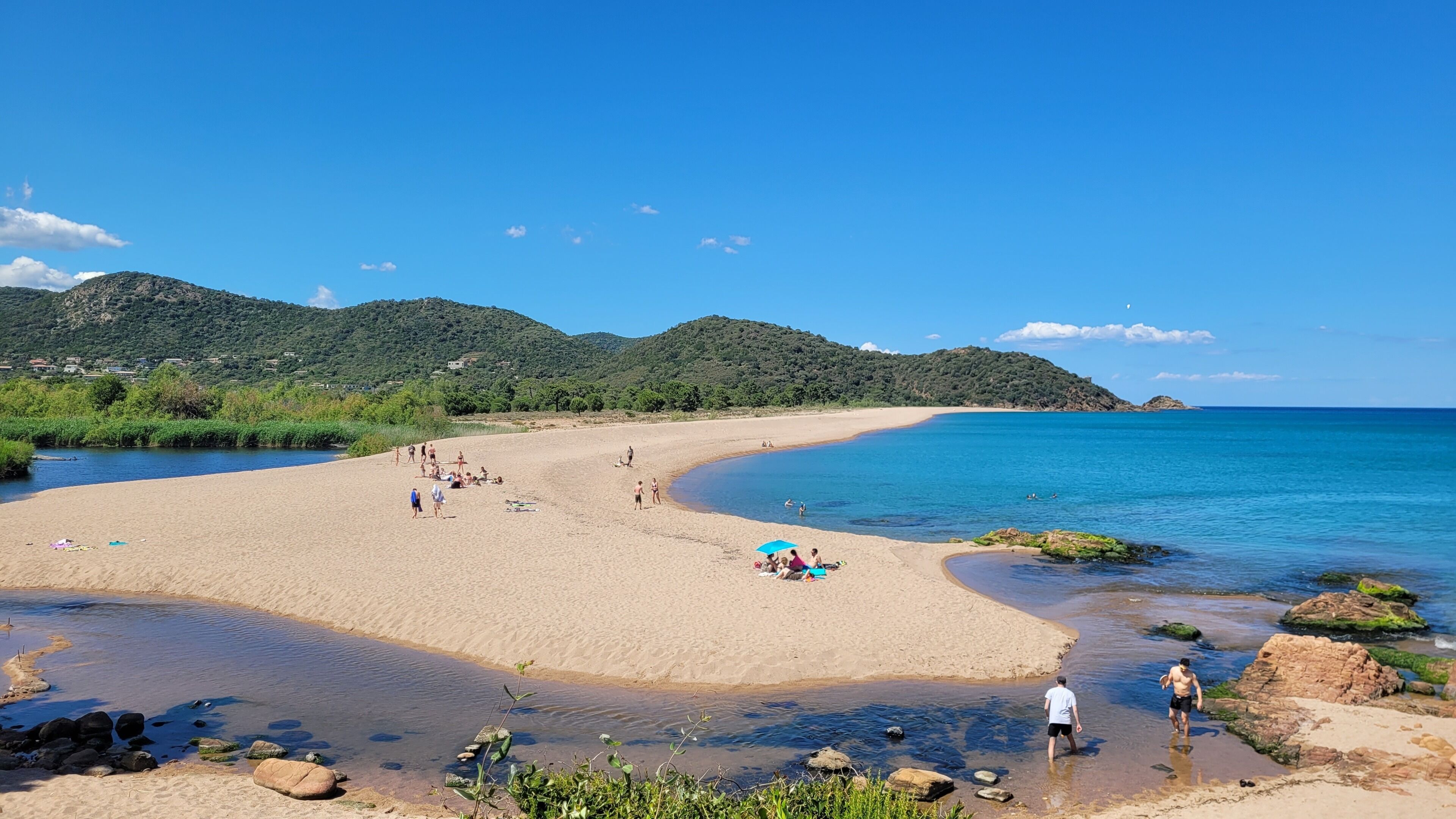 Une plage avec une eau cristalline, des gens qui prennent un bain de soleil et une montagne en arrière-plan.