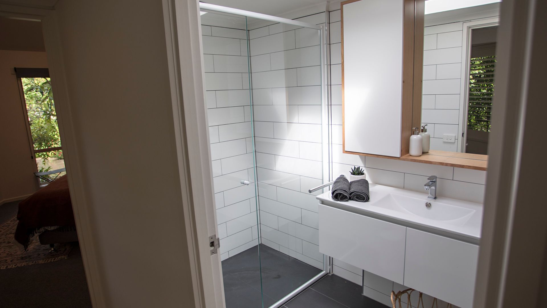 A modern bathroom with white tiles, a glass shower enclosure, a sink with a mirror, and a small potted plant.
