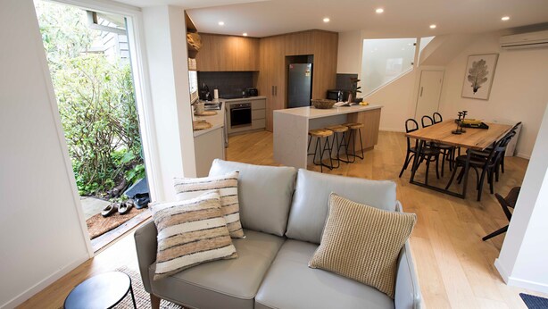 Ein modernes Wohnzimmer mit einer grauen Couch, einem Essbereich mit Holztisch und -stühlen, einer Küche mit Holzschränken und einem schwarzen Kühlschrank, und Blick auf Grünflächen.