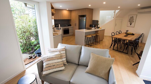 A modern living room with a grey sofa, a dining area with a wooden table and chairs, a kitchen with wooden cabinets and a black refrigerator, and a view of greenery outside.