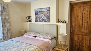A bedroom with a bed, a window with curtains, a bedside lamp, a wooden door, and a painting on the wall. - Saltburn Holidays Bluebell Cottage (Guisborough)
