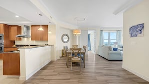Een moderne keuken met houten kastjes en een centraal eiland, een eetgedeelte met tafel en stoelen, en een woonkamer met een bank en een groot raam.