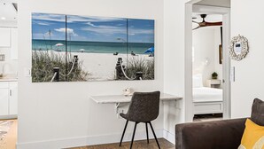 Une pièce comprenant un bureau, une chaise, un lit et une grande œuvre d’art sur le thème de la plage, accrochée au mur.