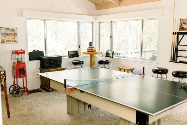 Una habitación con mesa de ping pong, dos sillas, una mesita con libros, un carrito rojo y una ventana que da a vistas de árboles.