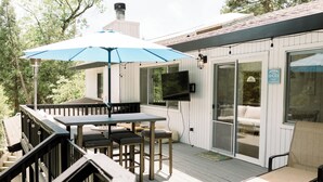 Una terraza cubierta al aire libre con una mesa y sillas, un televisor montado en la pared y un ventanal corredero que da a un espacio interior.