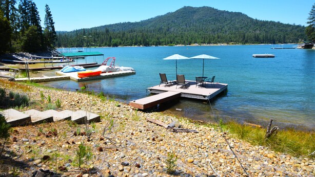 A lakeside dock with a boat, a seating area with umbrellas, and a forested hill in the background. - Lake Shore - Lake Front (Bass Lake)