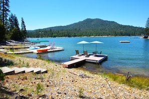 A lakeside dock with a boat, a seating area with umbrellas, and a forested hill in the background.